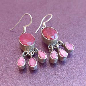 May include: A pair of silver dangle earrings with pink gemstone drops. The earrings feature a larger oval-shaped pink gemstone at the top and five smaller pink gemstone drops hanging below.