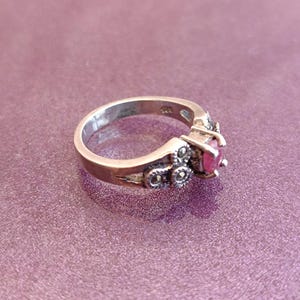 K&ouml;nnte beinhalten: Ein silberner Ring mit einem runden, leuchtend roten Edelstein. Der Ring hat dekorative Akzente und ist vor einem lila Hintergrund platziert. Ein Schmuckst&uuml;ck.