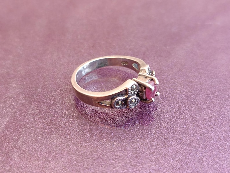 K&ouml;nnte beinhalten: Ein silberner Ring mit einem runden, leuchtend roten Edelstein. Der Ring hat dekorative Akzente und ist vor einem lila Hintergrund platziert. Ein Schmuckst&uuml;ck.