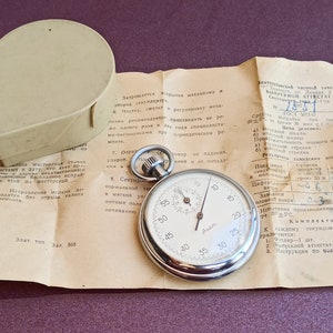 May include: A silver stopwatch with a white face and black markings. The stopwatch is in a box with a paper document that includes the text "ВИПУСКНОЙ АТТЕСТАТ" and "ГОСТ 072-61".