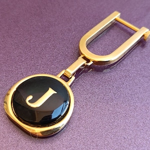 May include: Gold-tone key chain with a black circular charm featuring the letter "J" in gold.