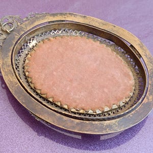 K&ouml;nnte beinhalten: Ein antiker, ovaler Rahmen mit einem verwitterten Gold-Finish. Der Rahmen hat ein inneres dekoratives Metallfiligran und einen rosa Samteinsatz. Das verzierte Design umfasst eine dekorative obere Schlaufe.