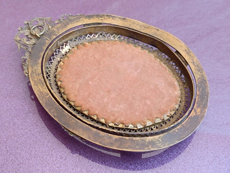 K&ouml;nnte beinhalten: Ein antiker, ovaler Rahmen mit einem verwitterten Gold-Finish. Der Rahmen hat ein inneres dekoratives Metallfiligran und einen rosa Samteinsatz. Das verzierte Design umfasst eine dekorative obere Schlaufe.