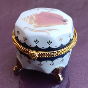 May include: A small white porcelain trinket box with a gold trim and a seashell design. The lid features a detailed seashell with a pink and brown color scheme. The box has three gold feet.