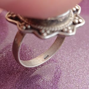 Op de afbeelding: Close-up van een zilveren ring met een bloemmotief. De ring heeft een gedetailleerde, sierlijke zetting met een centraal ontwerp en een band. De ring ligt op een paars oppervlak.