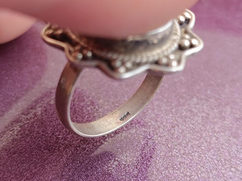 Op de afbeelding: Close-up van een zilveren ring met een bloemmotief. De ring heeft een gedetailleerde, sierlijke zetting met een centraal ontwerp en een band. De ring ligt op een paars oppervlak.