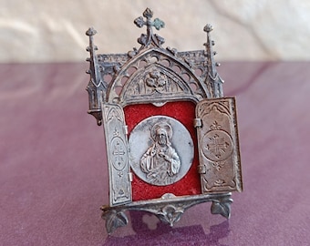 May include: A small, silver-colored, ornate, gothic-style reliquary with a red velvet interior and a silver medallion depicting Jesus Christ with his heart exposed.
