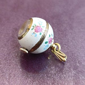 May include: A white, spherical pendant watch with a floral design in pink, blue, and green. The watch has gold-colored accents and a small crown. The pendant is attached to a gold-colored bail.