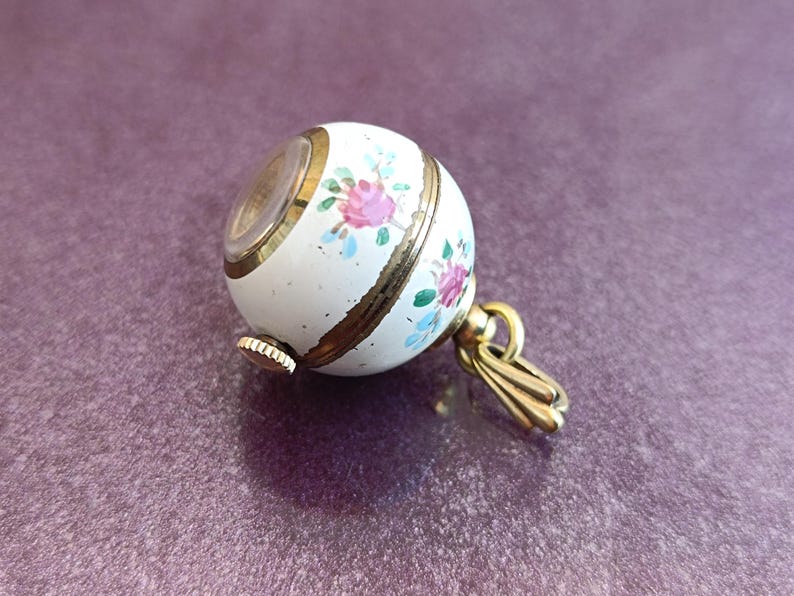 May include: A white, spherical pendant watch with a floral design in pink, blue, and green. The watch has gold-colored accents and a small crown. The pendant is attached to a gold-colored bail.
