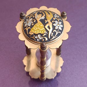 May include: A gold-toned hourglass with a decorative top featuring a black and gold design of two dancers and floral accents.