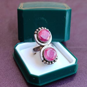Puede incluir: Un anillo de plata con dos grandes piedras preciosas rojas brillantes de corte ovalado. El anillo, de estilo vintage, se presenta en un joyero verde con interior blanco.