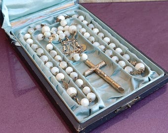 May include: An antique rosary featuring white beads and a silver cross, displayed in a vintage black box with a light blue satin lining. The rosary is a religious item.