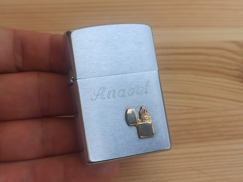 K&ouml;nnte beinhalten: Silberner Feuerzeug mit einer personalisierten Inschrift "Anabel" und einem kleinen goldenen Feuerzeugdesign.