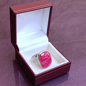 Op de afbeelding: Een zilveren ring met een grote, ovaalvormige, gefacetteerde, roze edelsteen wordt gepresenteerd in een open bordeauxrode sieradendoos met een witte satijnen binnenkant. De ring is het middelpunt.