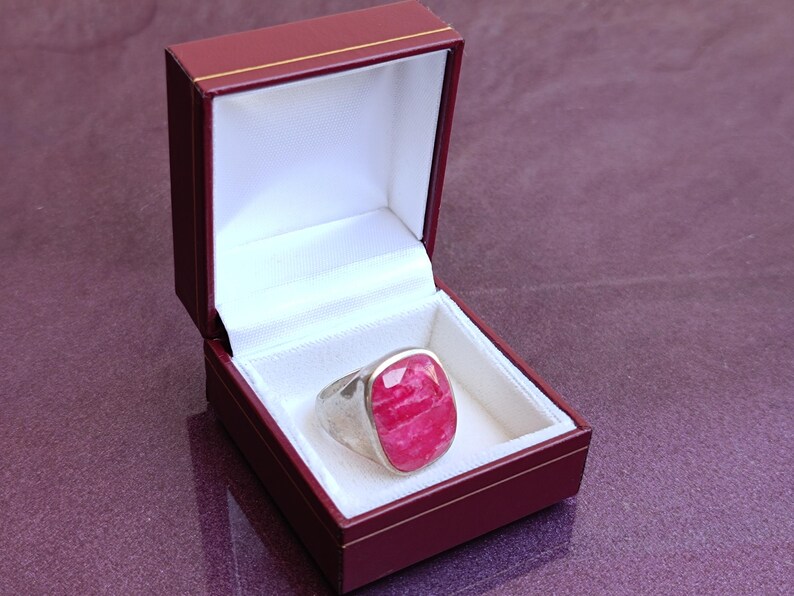 Op de afbeelding: Een zilveren ring met een grote, ovaalvormige, gefacetteerde, roze edelsteen wordt gepresenteerd in een open bordeauxrode sieradendoos met een witte satijnen binnenkant. De ring is het middelpunt.