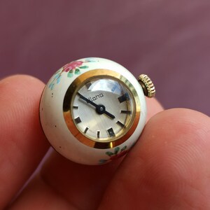 May include: A vintage, spherical watch with a white face and gold-tone accents. The watch face has black hour markers and hands. The watch casing is white with hand-painted floral designs in pink, blue, and green. The watch is labeled "Lana".