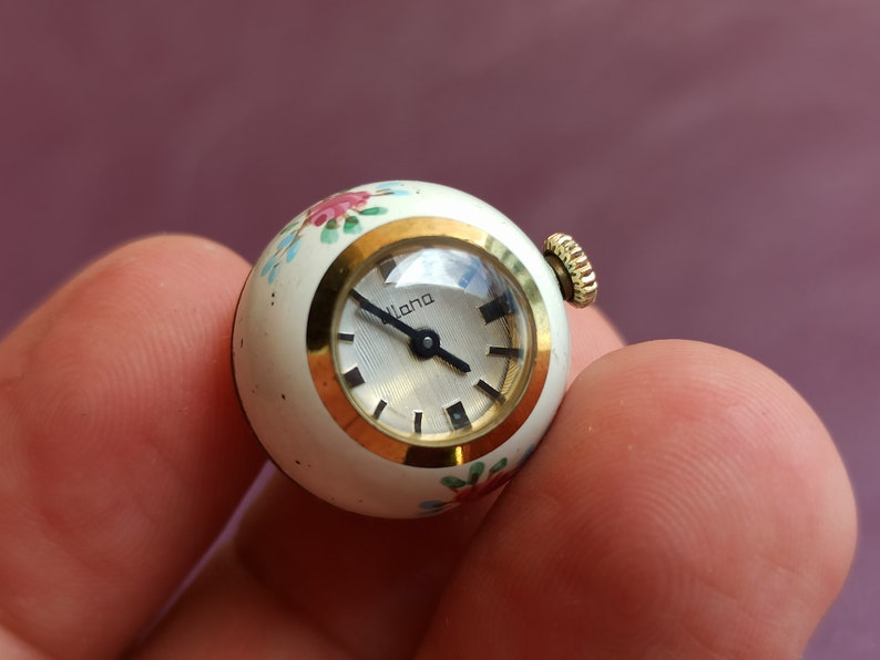 May include: A vintage, spherical watch with a white face and gold-tone accents. The watch face has black hour markers and hands. The watch casing is white with hand-painted floral designs in pink, blue, and green. The watch is labeled "Lana".