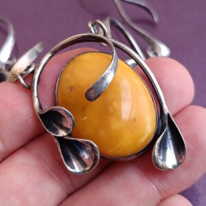May include: A silver pendant necklace with a large oval amber stone. The pendant features a stylized design with two leaf-like shapes on either side of the stone.