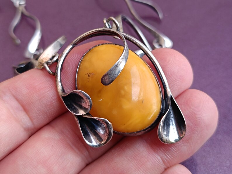 May include: A silver pendant necklace with a large oval amber stone. The pendant features a stylized design with two leaf-like shapes on either side of the stone.