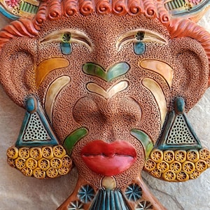 May include: A decorative ceramic wall mask featuring a textured brown face with vibrant red lips, green accents, and orange hair. The mask displays intricate geometric patterns, triangular earrings with yellow circular details, and blue eye highlights.