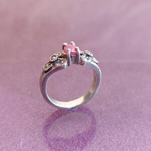 K&ouml;nnte beinhalten: Ein silberner Ring mit einem rosa Edelstein in der Mitte. Der Ring hat dekorative Verzierungen am Band und um den Edelstein herum. Der Ring ist vor einem lila Hintergrund platziert, der sein Design und seine Details hervorhebt.