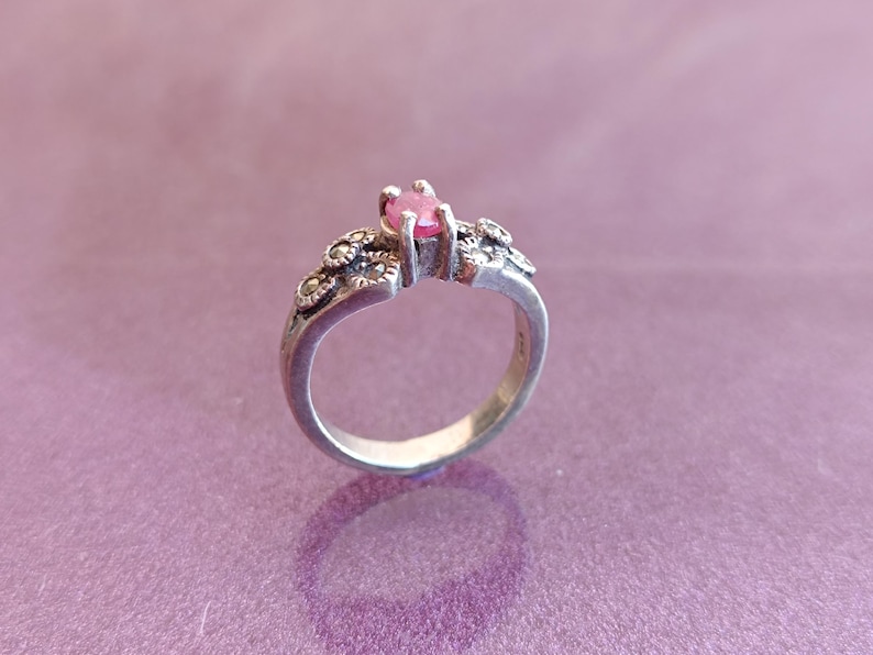 K&ouml;nnte beinhalten: Ein silberner Ring mit einem rosa Edelstein in der Mitte. Der Ring hat dekorative Verzierungen am Band und um den Edelstein herum. Der Ring ist vor einem lila Hintergrund platziert, der sein Design und seine Details hervorhebt.