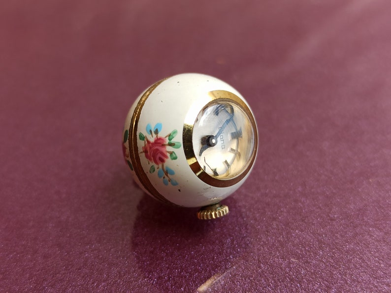 May include: A vintage, spherical watch with a cream-colored body and gold accents. The watch face is visible, and the body is decorated with a floral pattern in pink, blue, and green. The watch has a gold winding stem.
