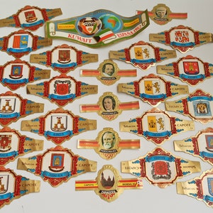 May include: A collection of colourful cigar band labels with gold trim. The labels feature various crests, coats of arms, and text in Spanish and Arabic. The labels are arranged in rows on a white surface.