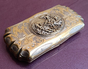 Può includere: Una scatola rettangolare in argento e oro con un design ornamentale. La scatola ha un pezzo centrale circolare rialzato con una scena di persone in un giardino.