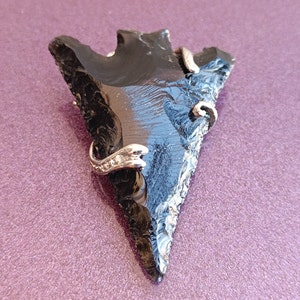 Op de afbeelding: Een zwarte obsidiaan pijlpunthanger met een zilverkleurig metalen accent. De pijl heeft een driehoekige vorm met een gekartelde rand. Het metalen accent is om de pijl gewikkeld en heeft kleine, heldere stenen.