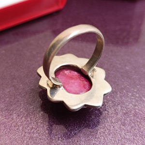 Op de afbeelding: Een zilveren ring met een grote, ovale, roze edelsteen gezet in een bloemvormige bezel. De ring wordt van onderaf bekeken, waarbij de zetting en de band zichtbaar zijn. De achtergrond is een paars oppervlak.