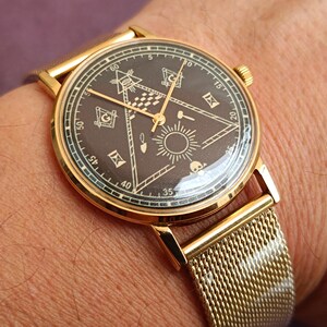 Könnte beinhalten: Eine goldfarbene Armbanduhr mit dunklem Zifferblatt, das Freimaurersymbole wie Zirkel, Winkel und Sonne zeigt. Die Uhr hat ein Mesh-Armband und ein goldenes Gehäuse. Die Uhr befindet sich an einem Handgelenk.