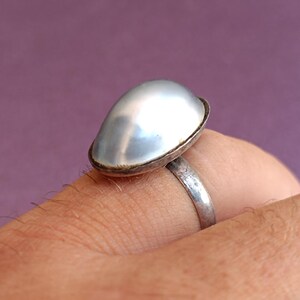 Puede incluir: Un anillo de plata con una piedra grande, nacarada y de forma ovalada. La piedra está engastada en un bisel de plata y se asienta sobre una fina banda de plata. El anillo se muestra en un dedo sobre un fondo púrpura.