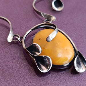 May include: A silver pendant necklace with a large, oval-shaped amber stone. The stone is surrounded by silver leaves and a silver swirl design.