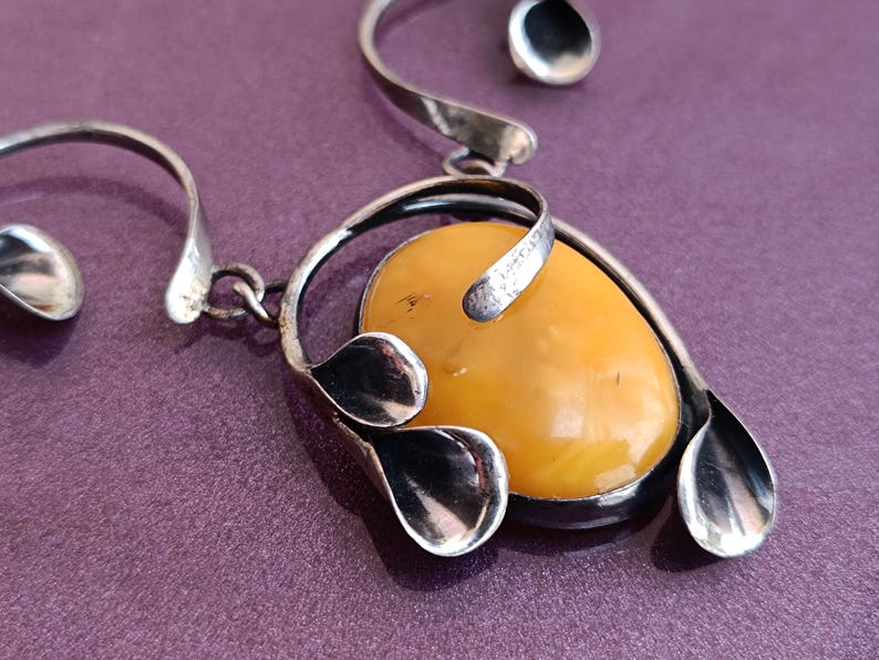 May include: A silver pendant necklace with a large, oval-shaped amber stone. The stone is surrounded by silver leaves and a silver swirl design.