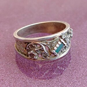 May include: A silver-toned metal ring featuring an openwork band with intricate scrollwork and floral-inspired patterns. A small, rectangular, teal-colored inlay is set into the side of the band, contrasting with the metallic texture of the ring.