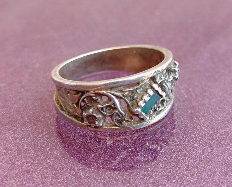 May include: A silver-toned metal ring featuring an openwork band with intricate scrollwork and floral-inspired patterns. A small, rectangular, teal-colored inlay is set into the side of the band, contrasting with the metallic texture of the ring.