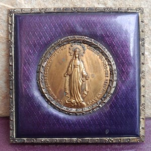 May include: A square religious icon featuring a gold-colored medallion of a saint against a purple background with a diamond pattern. The medallion is surrounded by a silver-colored frame and a decorative border.