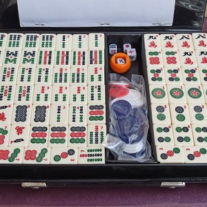 Może przedstawiać: Czarna walizka transportowa z białym zestawem mahjong w środku. Zestaw zawiera płytki z chińskimi znakami, liczbami i symbolami, a także czerwone i białe kostki oraz zestaw żeton&oacute;w.