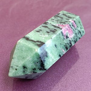 May include: A polished, rectangular gemstone with a faceted end. The stone is primarily green with black and pink inclusions. The gemstone is displayed on a purple surface, showcasing its natural colors and textures.