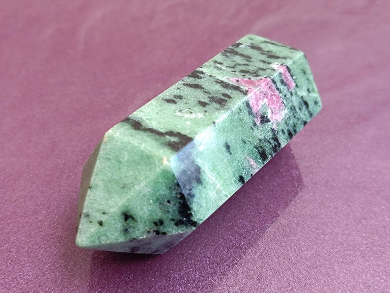 May include: A polished, rectangular gemstone with a faceted end. The stone is primarily green with black and pink inclusions. The gemstone is displayed on a purple surface, showcasing its natural colors and textures.