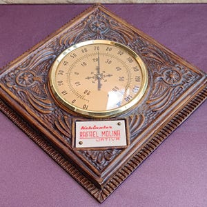 May include: A vintage wooden thermometer with a brass-colored face and a decorative, carved frame. The thermometer displays temperature readings in Fahrenheit. A small plaque at the bottom reads "Kehinator RAFAEL MOLINA JATIVA."