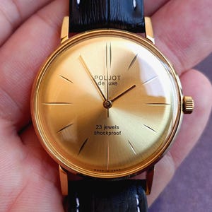 Op de afbeelding: Een goudkleurig analoog polshorloge met een ronde wijzerplaat, een goudkleurige sunburst-achtergrond, dunne gouden uurmarkeringen en gouden wijzers. De wijzerplaat bevat de tekst Poljot de luxe, 23 jewels, Shockproof en Made in USSR. Zwarte leren band.