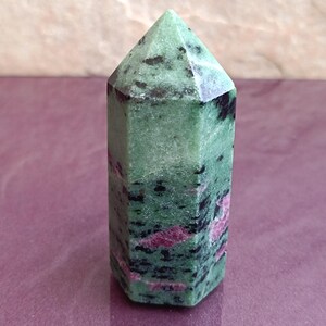 May include: A green and black zoisite crystal point with red ruby inclusions. The polished gemstone has a hexagonal base and a pointed top. The crystal is approximately 4 inches tall, perfect for display or collection.