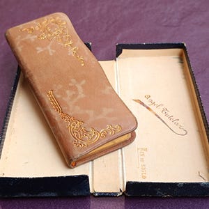 Könnte beinhalten: Ein braunes, ledergebundenes Tagebuch mit goldenen Prägungen und dem Text "Angel Tutelar" und "Fin de Siglo" auf dem Einband. Das Tagebuch ist geöffnet und liegt auf einer schwarzen Schachtel.