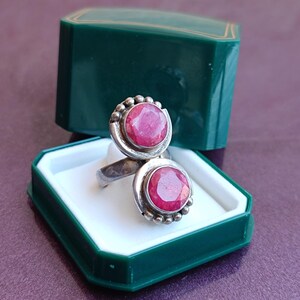 Puede incluir: Anillo de plata con dos grandes piedras preciosas redondas de color rojo brillante. El anillo se muestra en una caja de joyer&iacute;a verde con interior blanco. Las piedras preciosas est&aacute;n engastadas en un engaste de plata decorativo.
