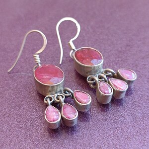 May include: Silver dangle earrings with pink gemstone drops. The earrings feature a larger oval-shaped pink gemstone at the top and smaller teardrop-shaped pink gemstones dangling below.