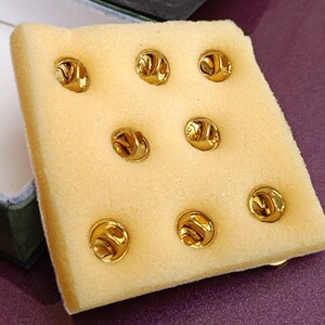 May include: A close-up of a white foam sheet with eight gold-colored metal pin backs. The pin backs are arranged in a grid pattern.