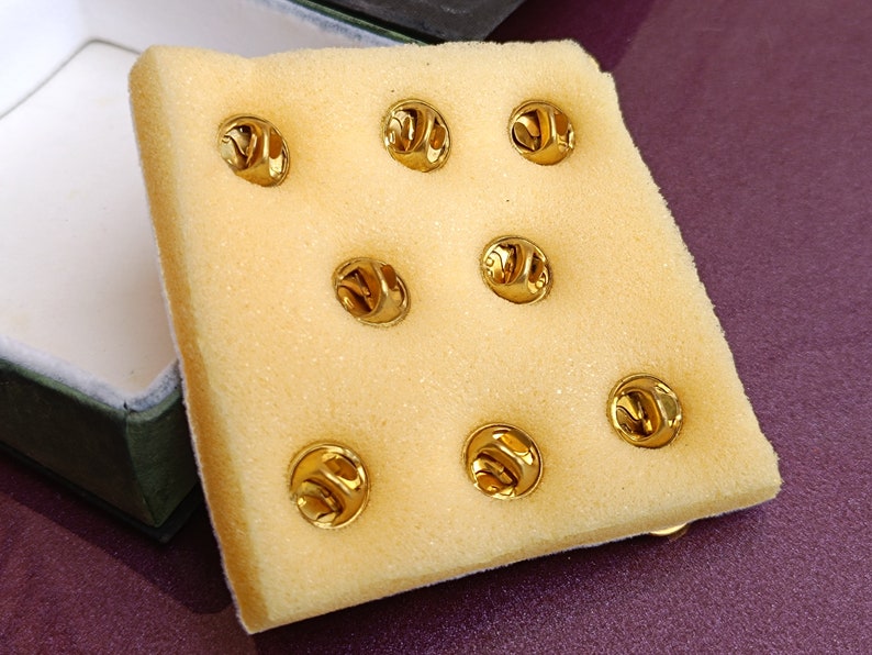 May include: A close-up of a white foam sheet with eight gold-colored metal pin backs. The pin backs are arranged in a grid pattern.