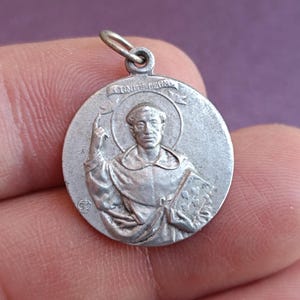 May include: A silver-coloured round pendant depicting a religious figure with a halo, holding a book and raising a hand. The pendant has the inscription "UNITÉ REINE" on a banner. It has a small loop at the top for a chain.